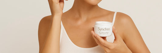 A woman with smooth, radiant skin applies Synchro Regulating Face Care cream to her cheek using a white spatula. She is wearing a white tank top and smiling gently, holding the product jar in her other hand against a soft, neutral background.